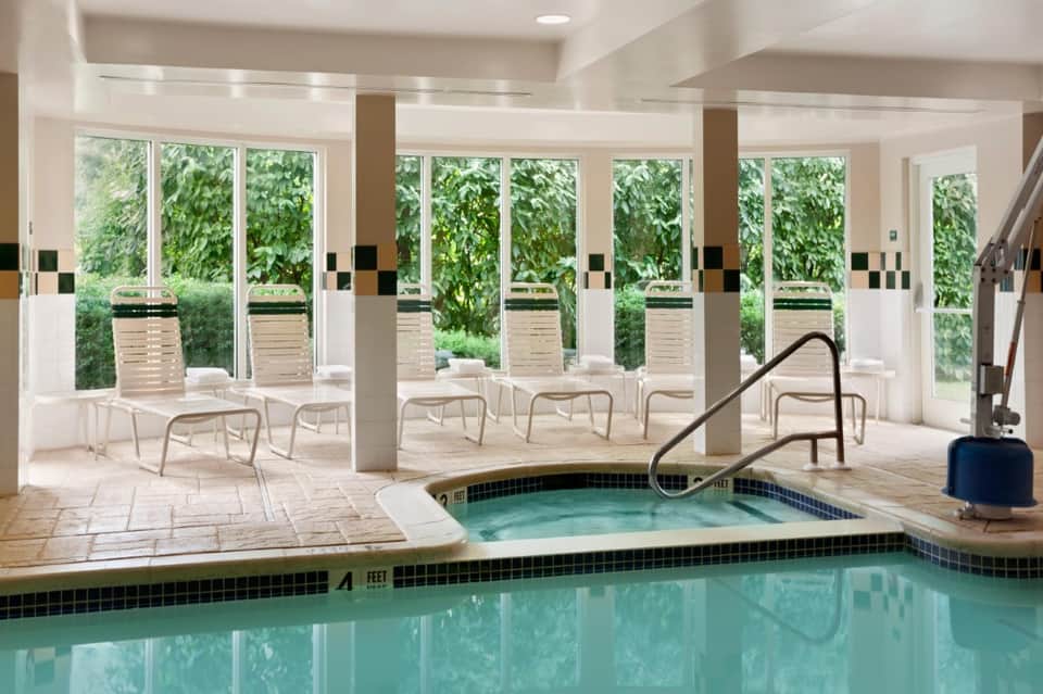 Indoor pool area with hot tub, lounge chairs, floor-to-ceiling windows overlooking garden, and spa facilities