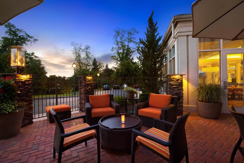 Twilight rooftop patio with wicker seating, orange cushions, fire pit, stone pillars, and lit building facade