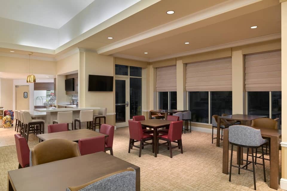 Hotel common area with multiple dining tables, burgundy chairs, bar, large windows, and bright ceiling lighting