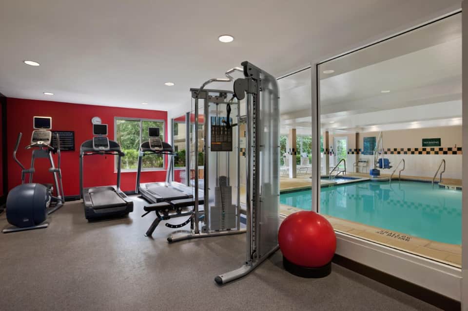 Fitness center with red wall, cardio equipment, weight machines, and glass partition overlooking indoor pool