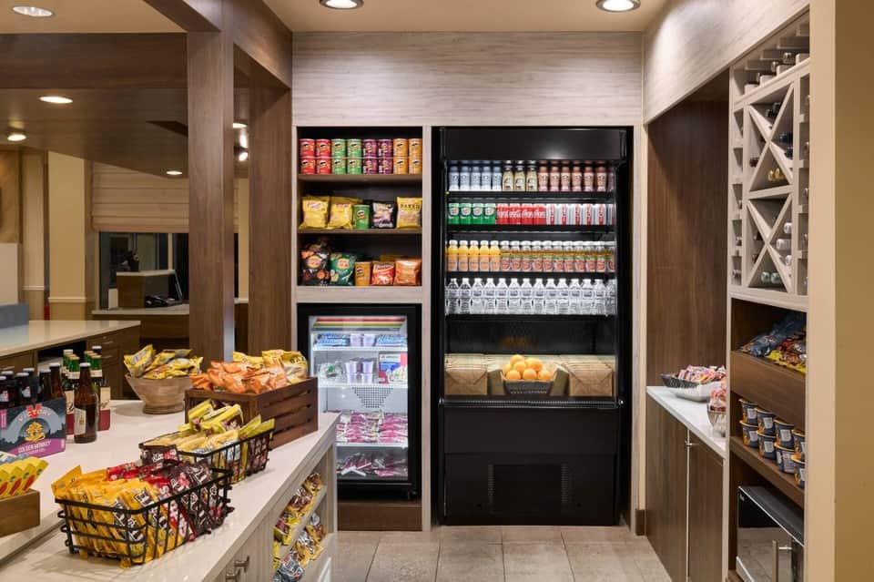 Modern hotel snack bar with refrigerated beverage display, shelving, and colorful packaged snacks