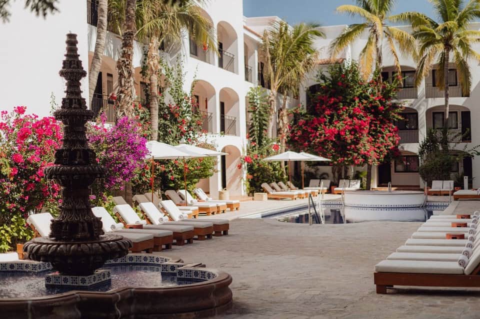 Luxury resort courtyard with white arched architecture, vibrant bougainvillea flowers, lounge chairs, and decorative fountain.