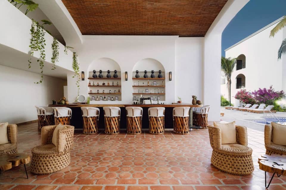 Outdoor bar with woven wicker bar stools, arched shelving displaying bottles, and resort courtyard views.