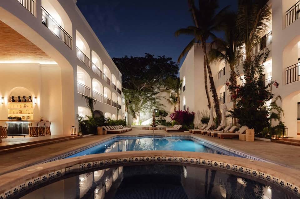 Nighttime courtyard pool with illuminated white building facades, palm trees, and decorative tile borders.