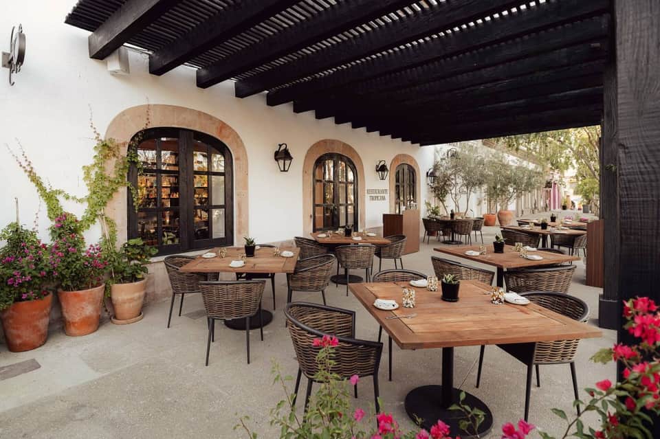 Open-air restaurant patio with wicker chairs, wooden tables, arched doorways, and pink flowering plants.