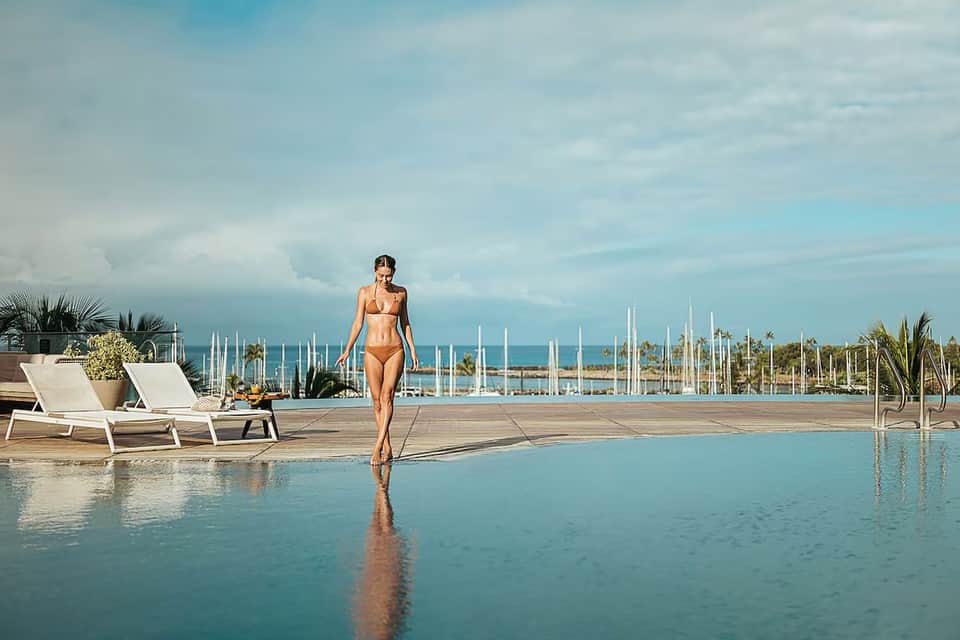 Woman walking along infinity pool edge with marina and sailing boats in background