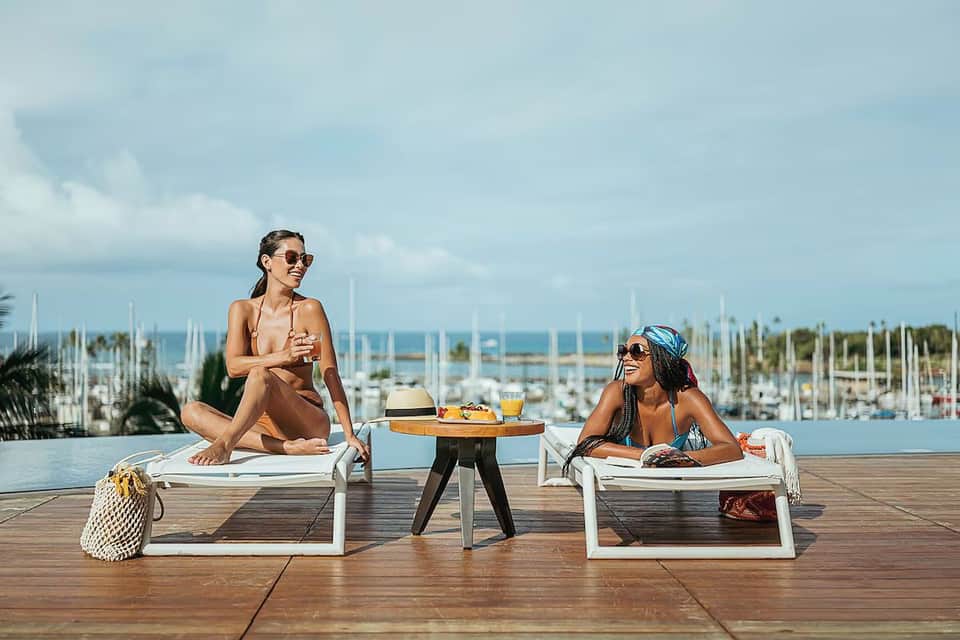 Two women relaxing on lounge chairs by infinity pool overlooking marina and sailboats