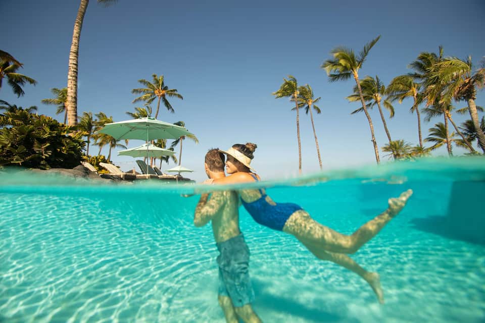 Split underwater view of two people in turquoise lagoon with palm trees and cabana on shore