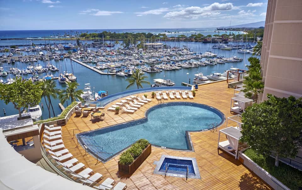 Resort pool deck overlooking marina filled with sailboats, featuring lounge chairs and waterfront high-rise building