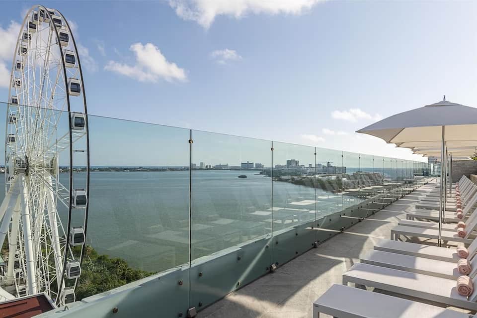 Rooftop lounge with white loungers, umbrellas, glass railing, and Ferris wheel in distance