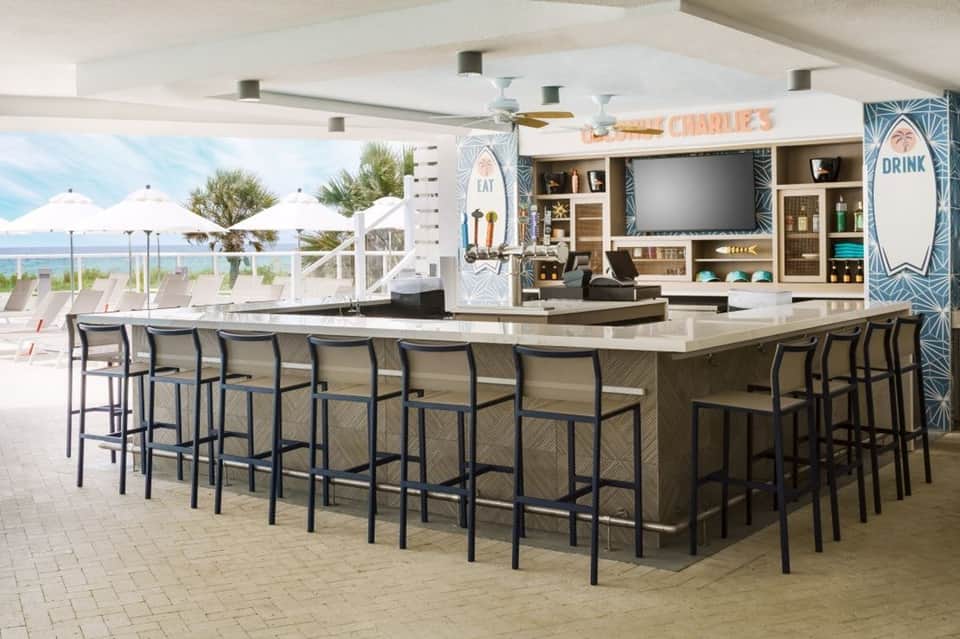 Beachfront bar and lounge area with ocean view, white umbrellas, and modern shelving displaying bottles and decor