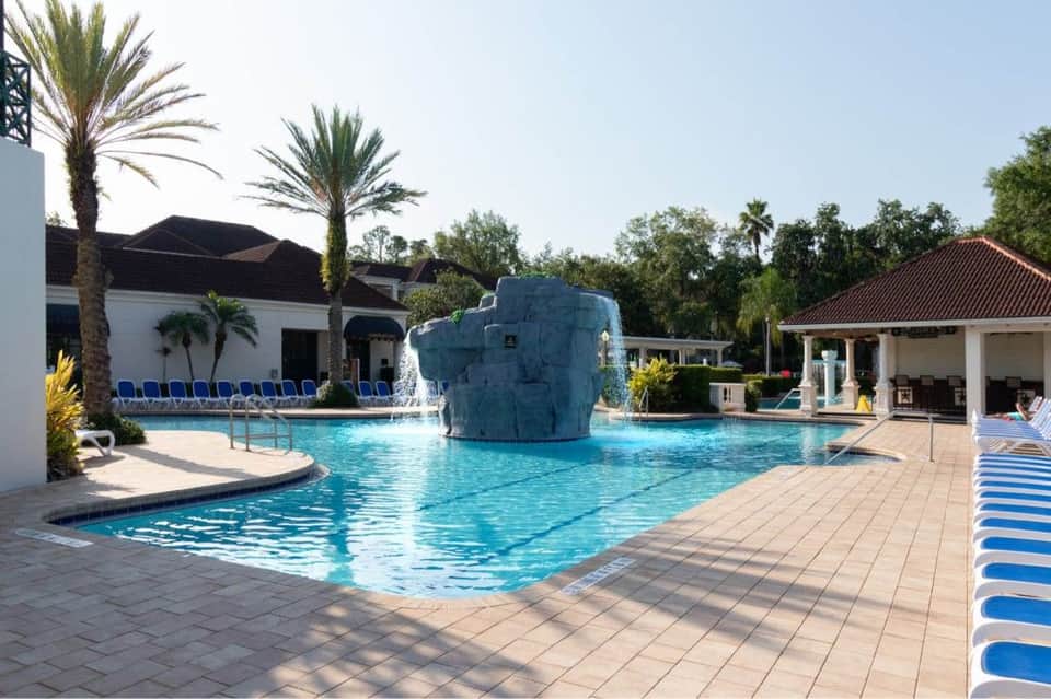 Resort pool with rock waterfall feature, palm trees, white buildings, and covered pavilion