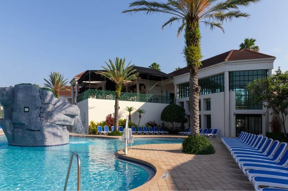 Large resort pool with sculptural rock feature, palm trees, covered structures, and blue loungers