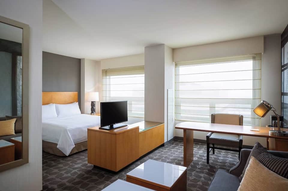 Modern hotel suite with bed, work desk, TV, and ocean view windows with sheer curtains