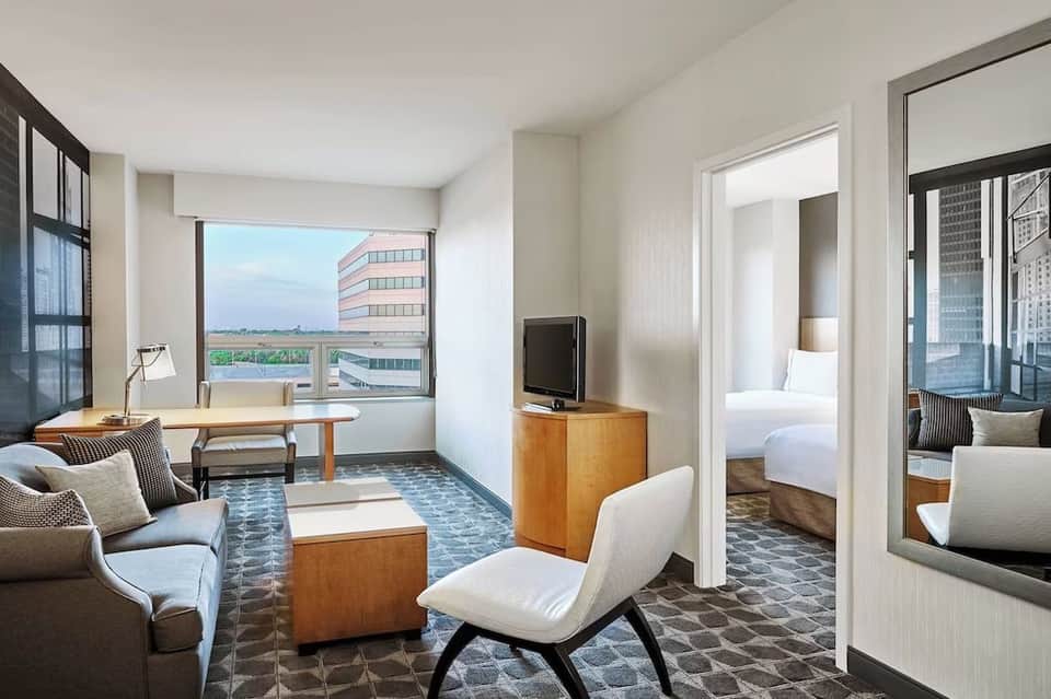 Hotel suite living area with city view, work desk, white modern furniture, and open bedroom doorway