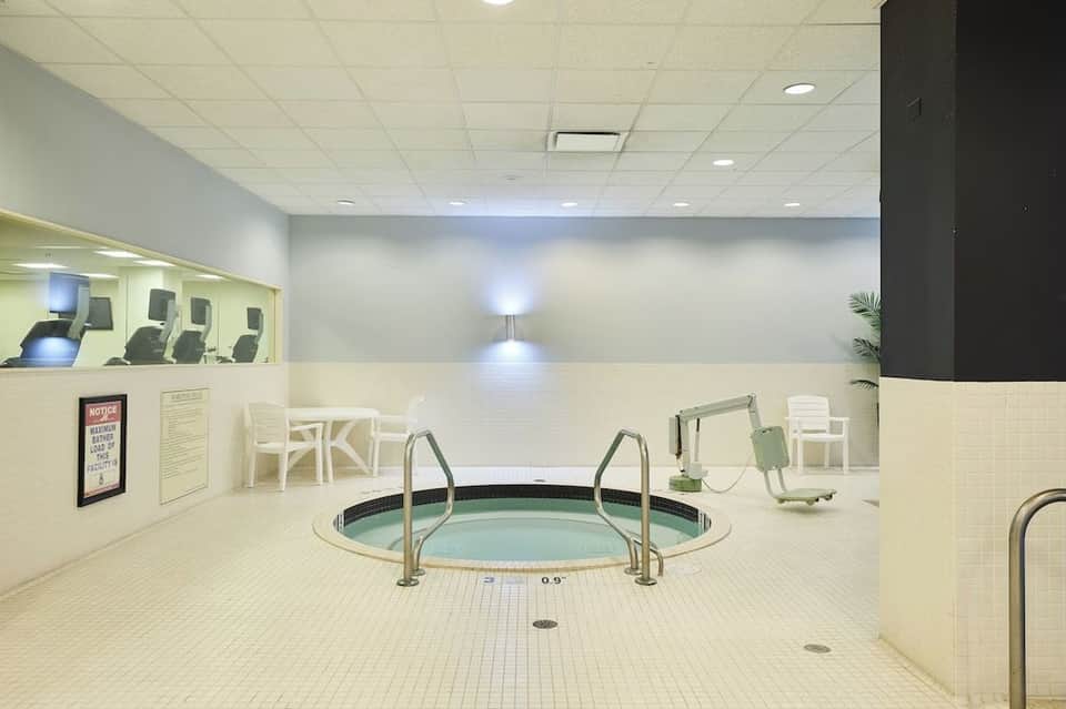 Indoor hot tub with metal railings in a cream tile spa room with fitness equipment visible in background