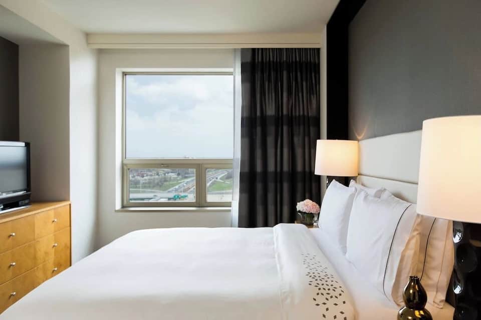 Hotel bedroom with large window overlooking cityscape, queen bed with white bedding, wood dresser, and reading chair