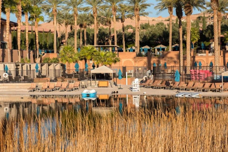 Waterfront resort with sandy beach, palm trees, lounge chairs, and multi-story buildings
