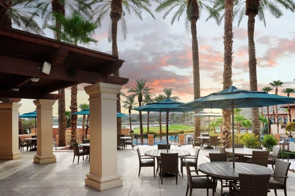 Covered patio with columns, dining tables, umbrellas, and palm trees at sunset