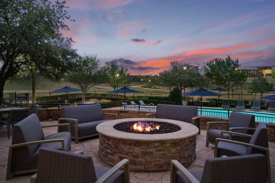 Outdoor fire pit lounge area at dusk with blue umbrellas, seating, and illuminated pool in background