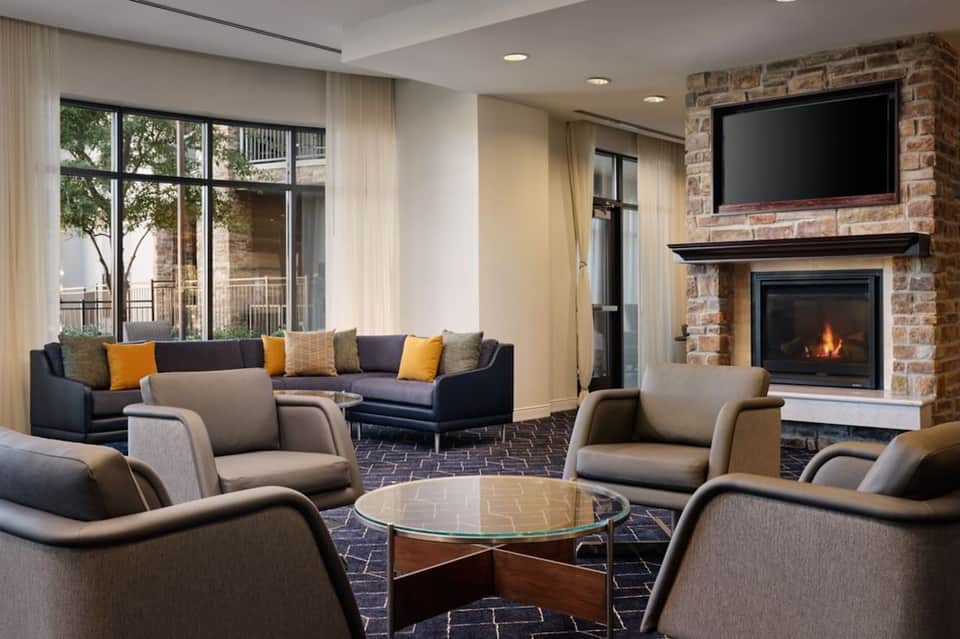 Modern hotel lobby with fireplace, brick accent wall, TV, comfortable seating, and large windows