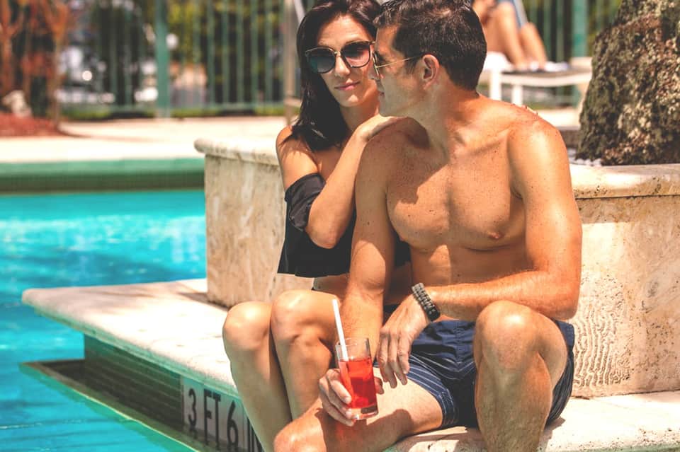 Couple relaxing by bright turquoise pool, man holding red beverage while sitting poolside