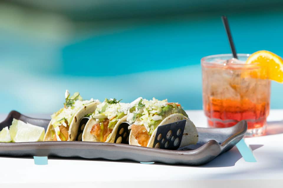 Fish tacos with crispy shells and fresh garnish beside a tropical cocktail by the pool