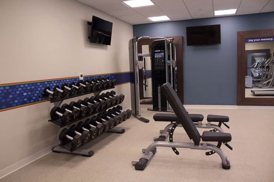 Hotel fitness center with dumbbell rack, weight bench, cable machine, and flat-screen TV