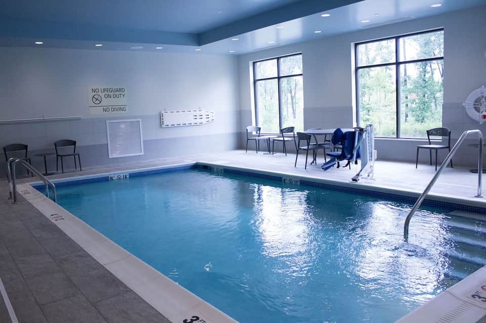 Indoor swimming pool with safety signage, chairs, large windows showing forest views, and teal ceiling