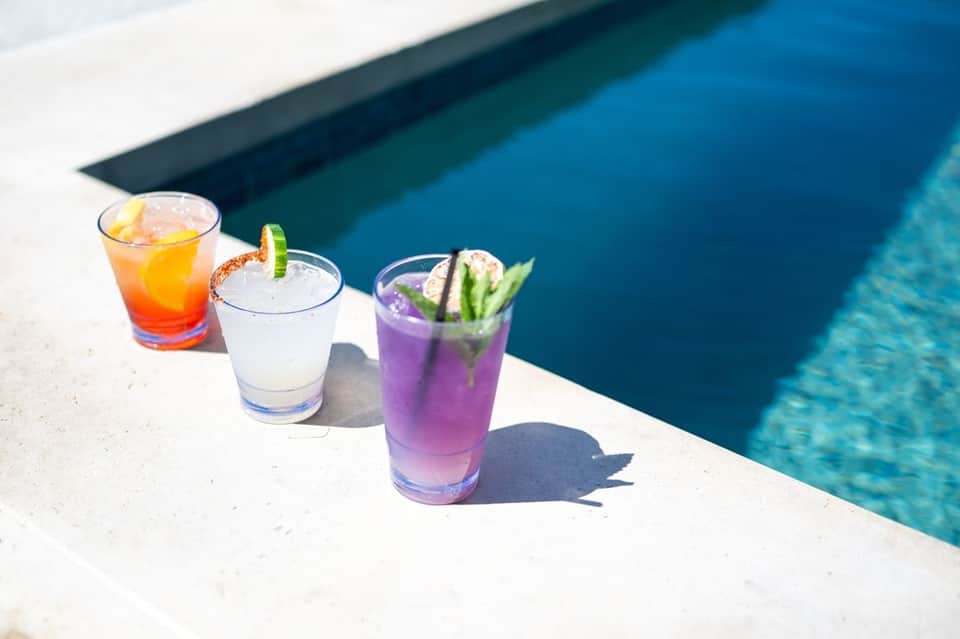 Three colorful cocktails on a white pool deck beside a turquoise swimming pool