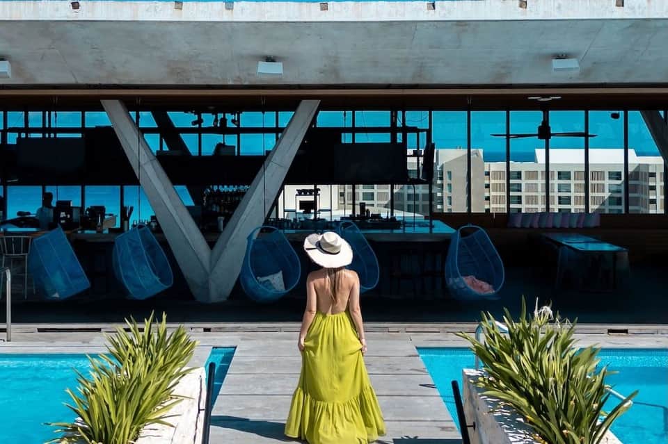 Woman in yellow dress standing poolside facing modern lounge with ocean views and large windows