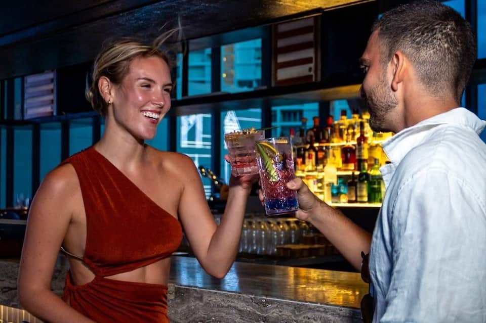 Couple clinking colorful cocktails at an upscale rooftop bar with city lights