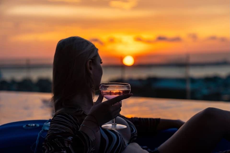 Woman holding wine glass watching golden sunset over waterfront from resort