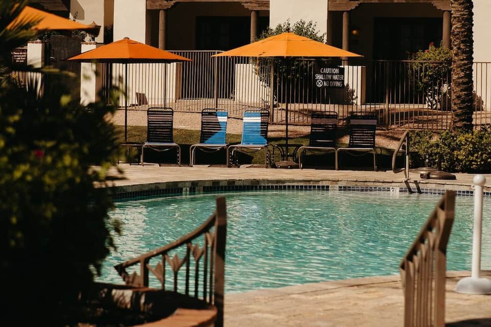 Resort pool with orange umbrellas, lounge chairs, white building, and shallow water warning sign