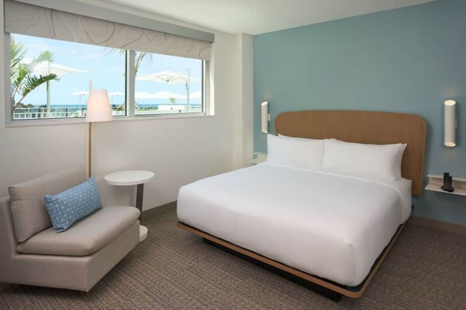 Hotel bedroom with light blue walls, wooden headboard, and window overlooking beach