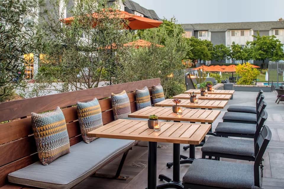 Rooftop terrace with wooden dining tables, patterned cushions, orange umbrellas, and olive trees overlooking residential buildings