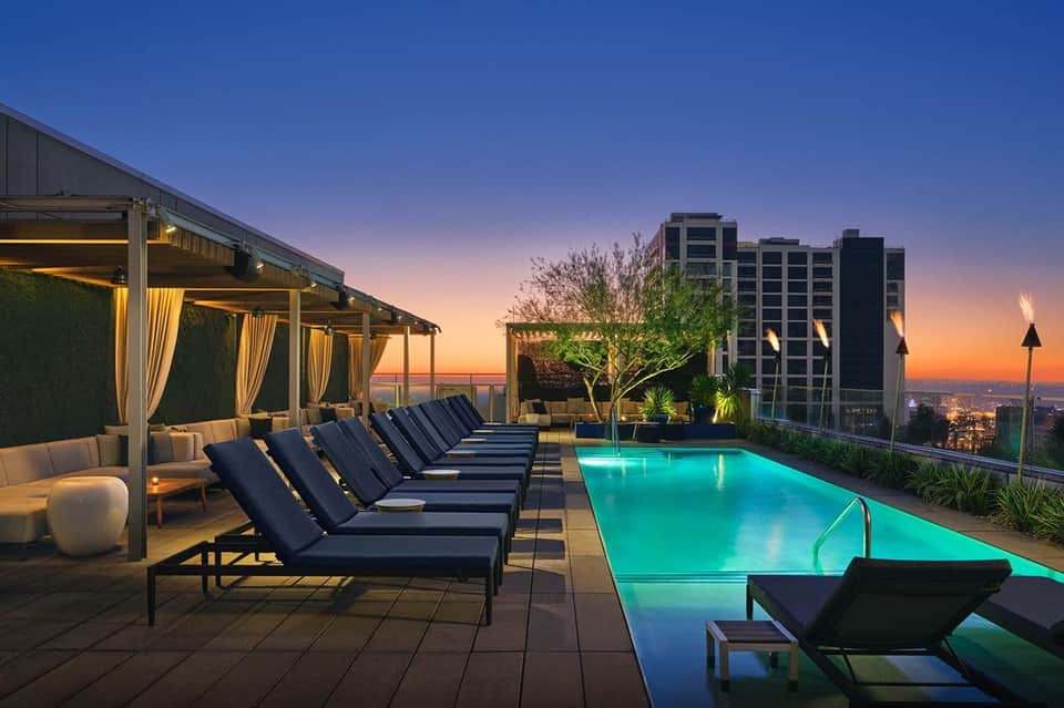 Rooftop pool at sunset with lounge chairs, pergola seating, glowing blue water, and city lights below