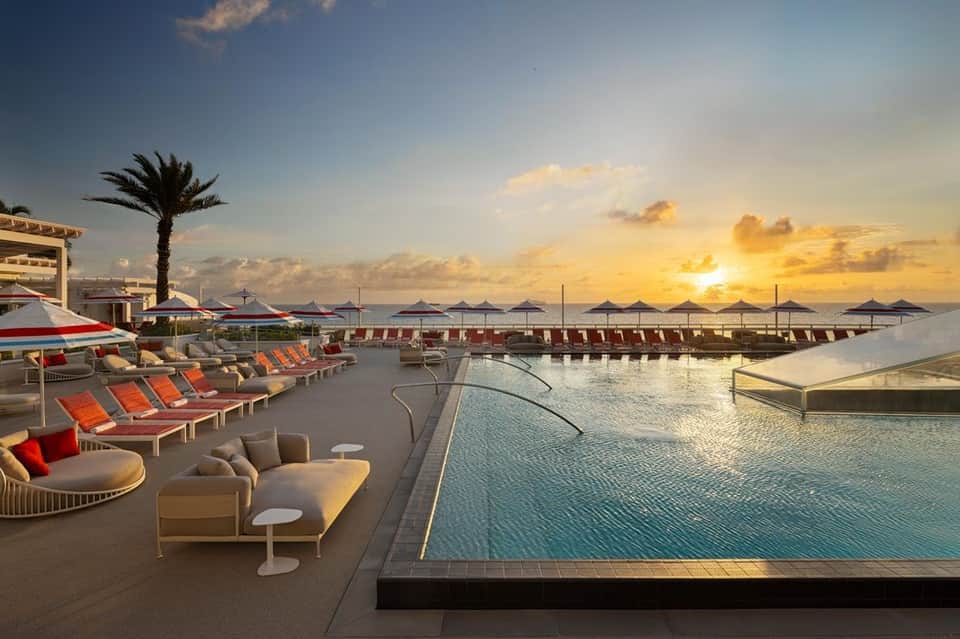 Luxury resort pool deck at sunset with red lounge chairs, umbrellas, and ocean view with palm trees