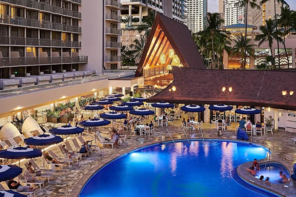 Twilight view of large resort pool with blue umbrellas, dining area, modern buildings, and lit architectural features