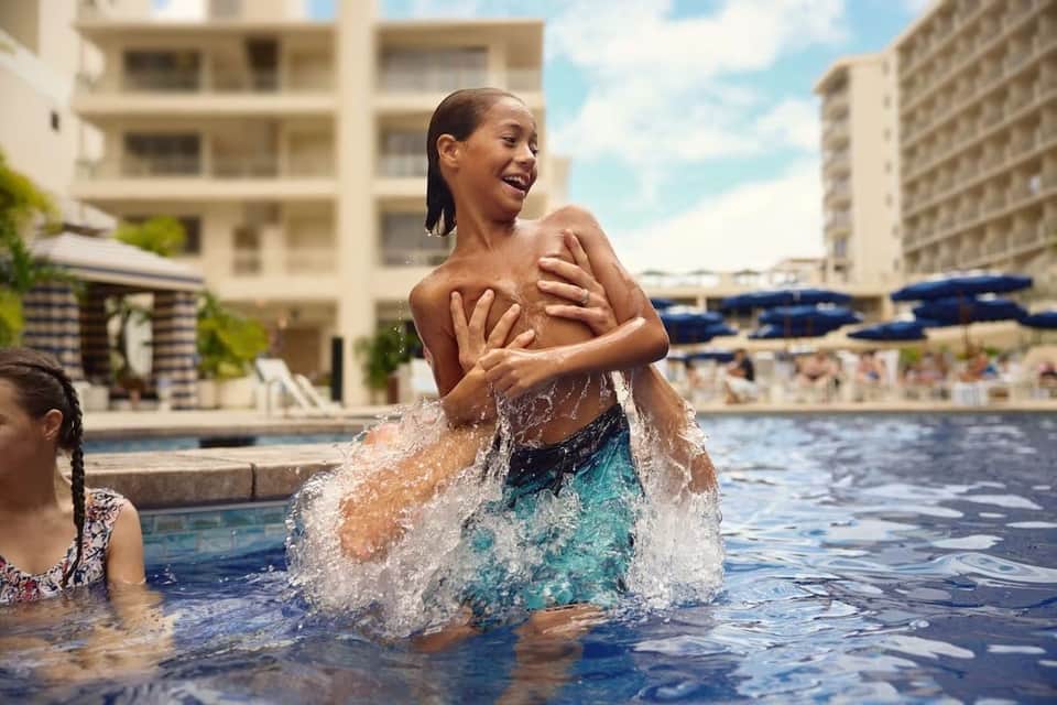 Young boy playing in resort pool with water splashing, surrounded by modern apartment buildings
