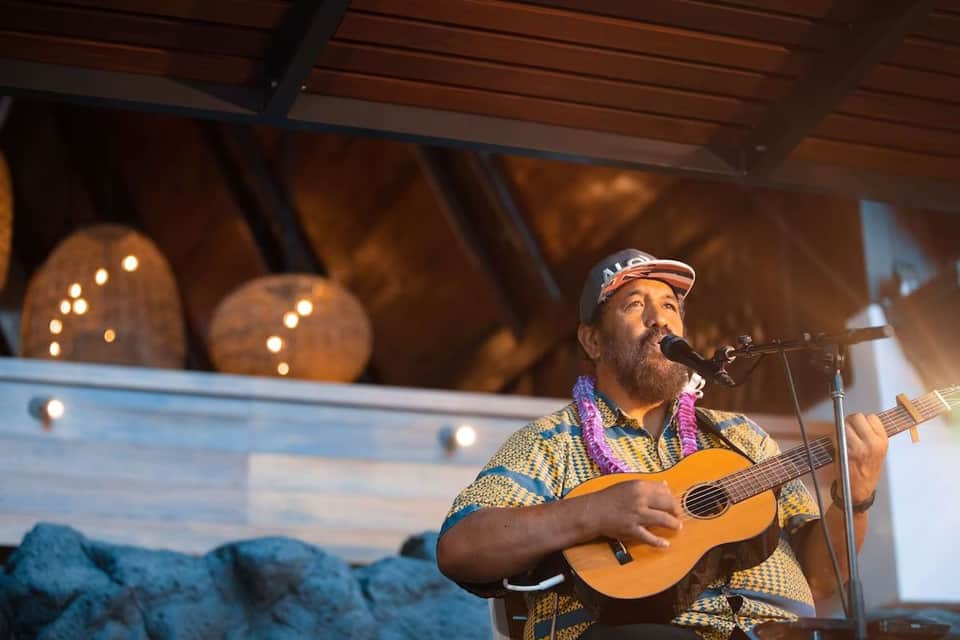 Male musician performing with guitar on indoor stage wearing colorful patterned shirt and lei