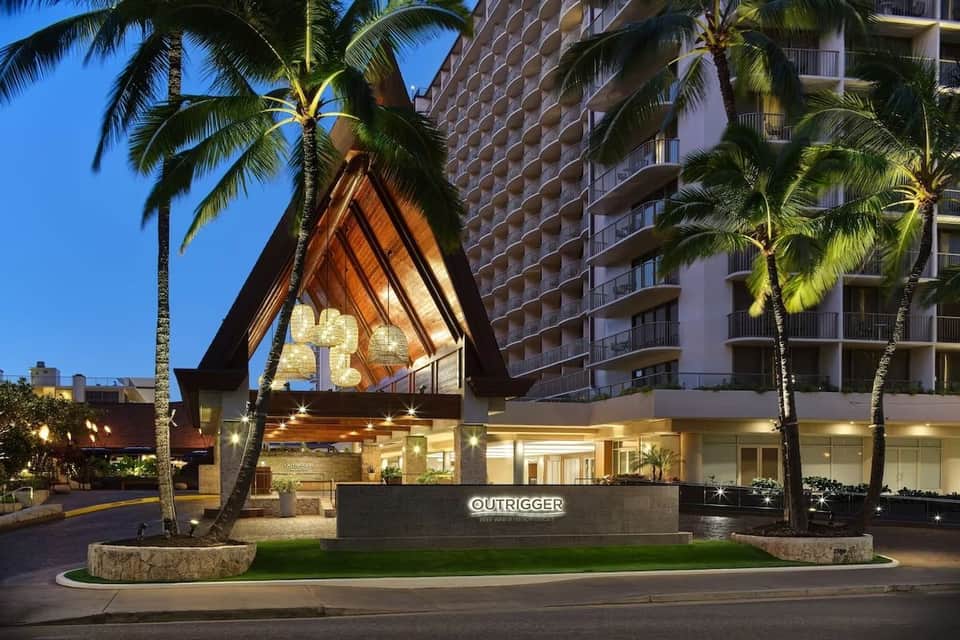 Modern resort entrance at dusk with distinctive A-frame architectural feature, illuminated signage, palm trees, and tall building