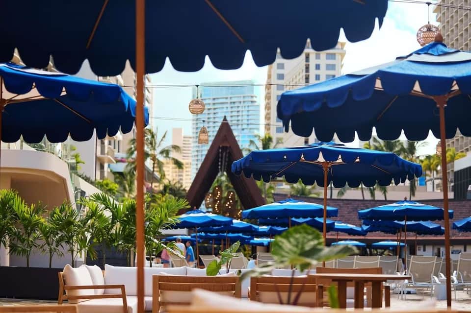 Resort pool deck with blue scalloped umbrellas, wooden loungers, and high-rise buildings