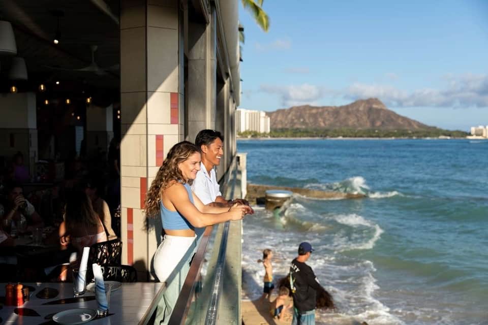 Beachfront bar with couple viewing ocean, Diamond Head mountain, and surfers in water