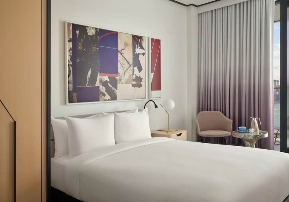 Modern hotel bedroom with white bedding, purple curtains, city skyline view, and wall art