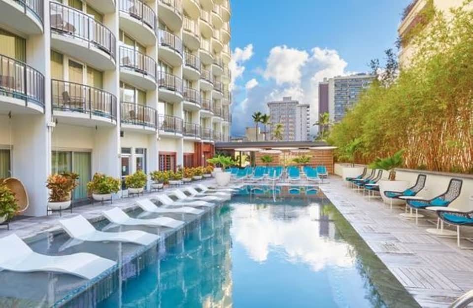 Modern hotel pool with blue loungers, white umbrellas, and multi-story building facade