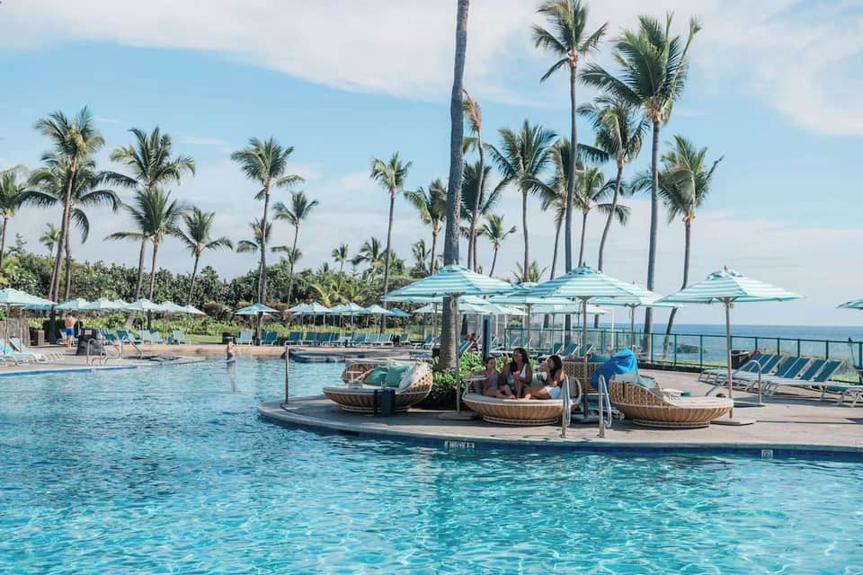 Resort pool with turquoise water, floating lounges, palm trees, and ocean view
