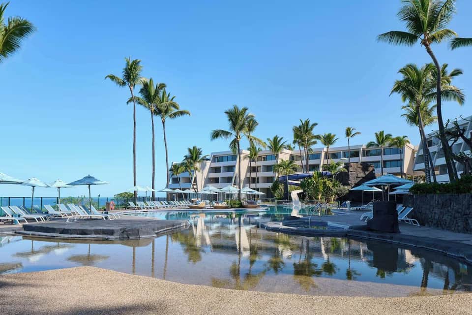 Curved resort pool with shallow area, lounge chairs, umbrellas, and beachfront hotel building