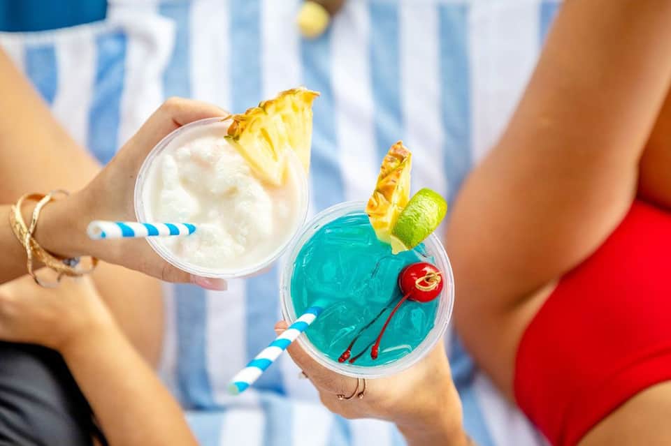 Two tropical cocktails with pineapple garnish and blue striped straws held by guests