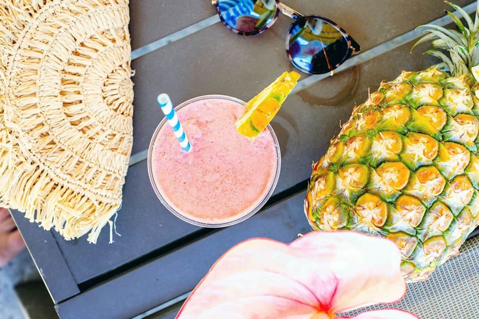 Tropical beach drink setup with pink smoothie, pineapple, straw hat, and sunglasses